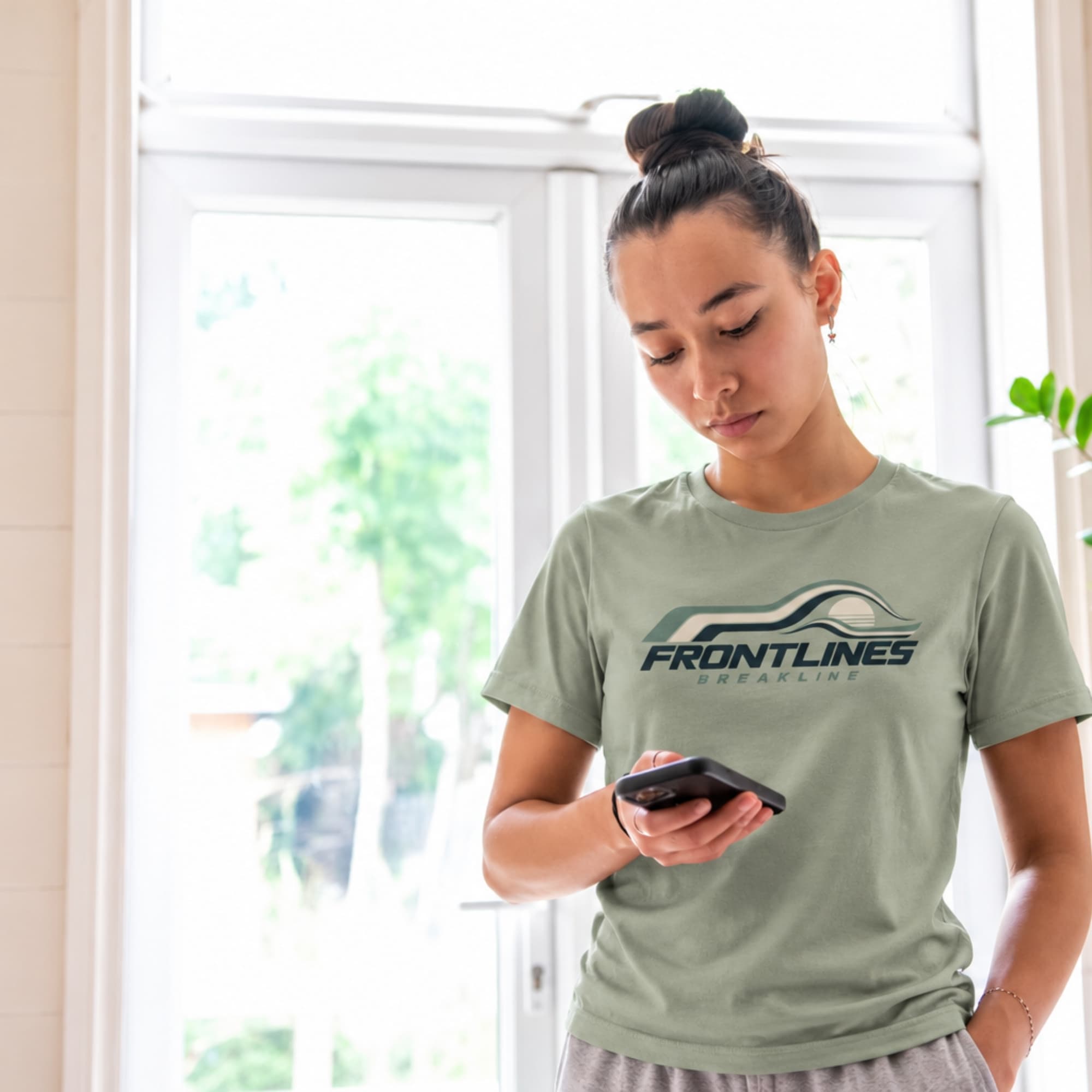A woman wearing a sage Breakline graphic tee by a window