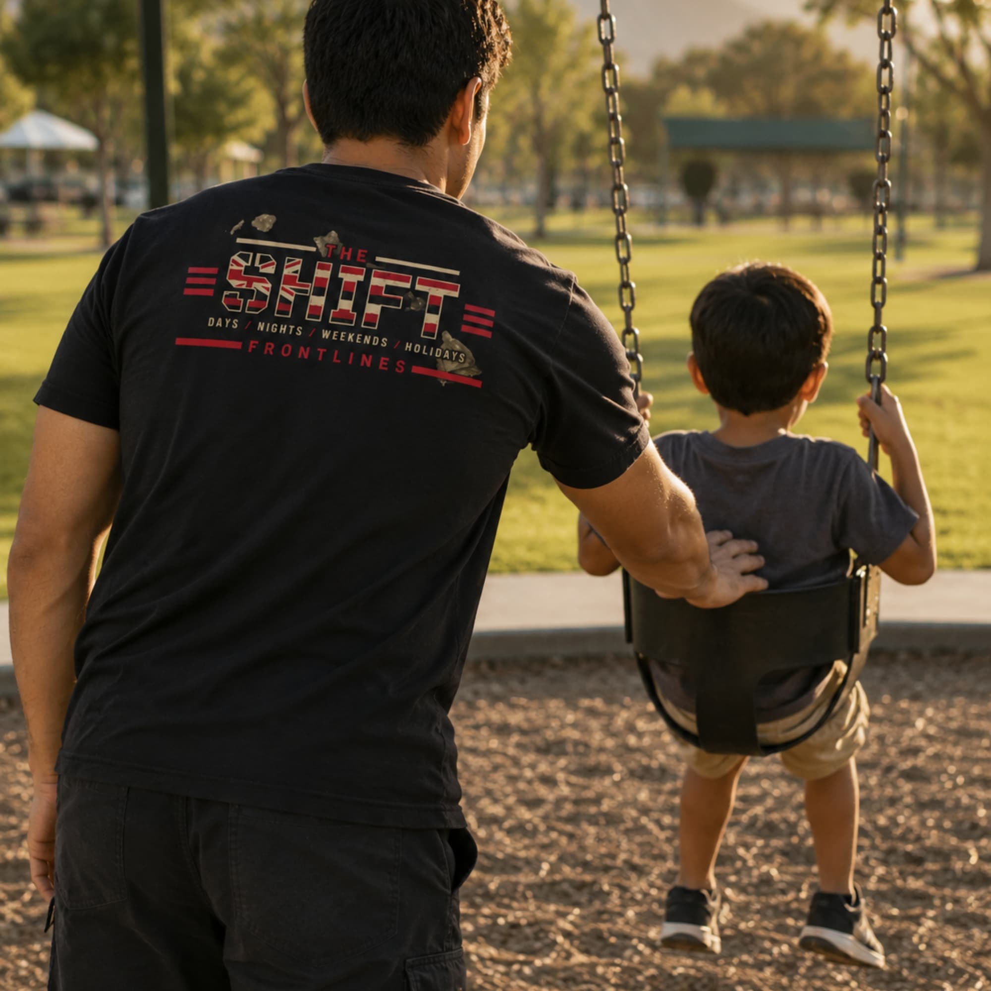 A man wearing a Frontlines Shift tee pushing a child on a swing